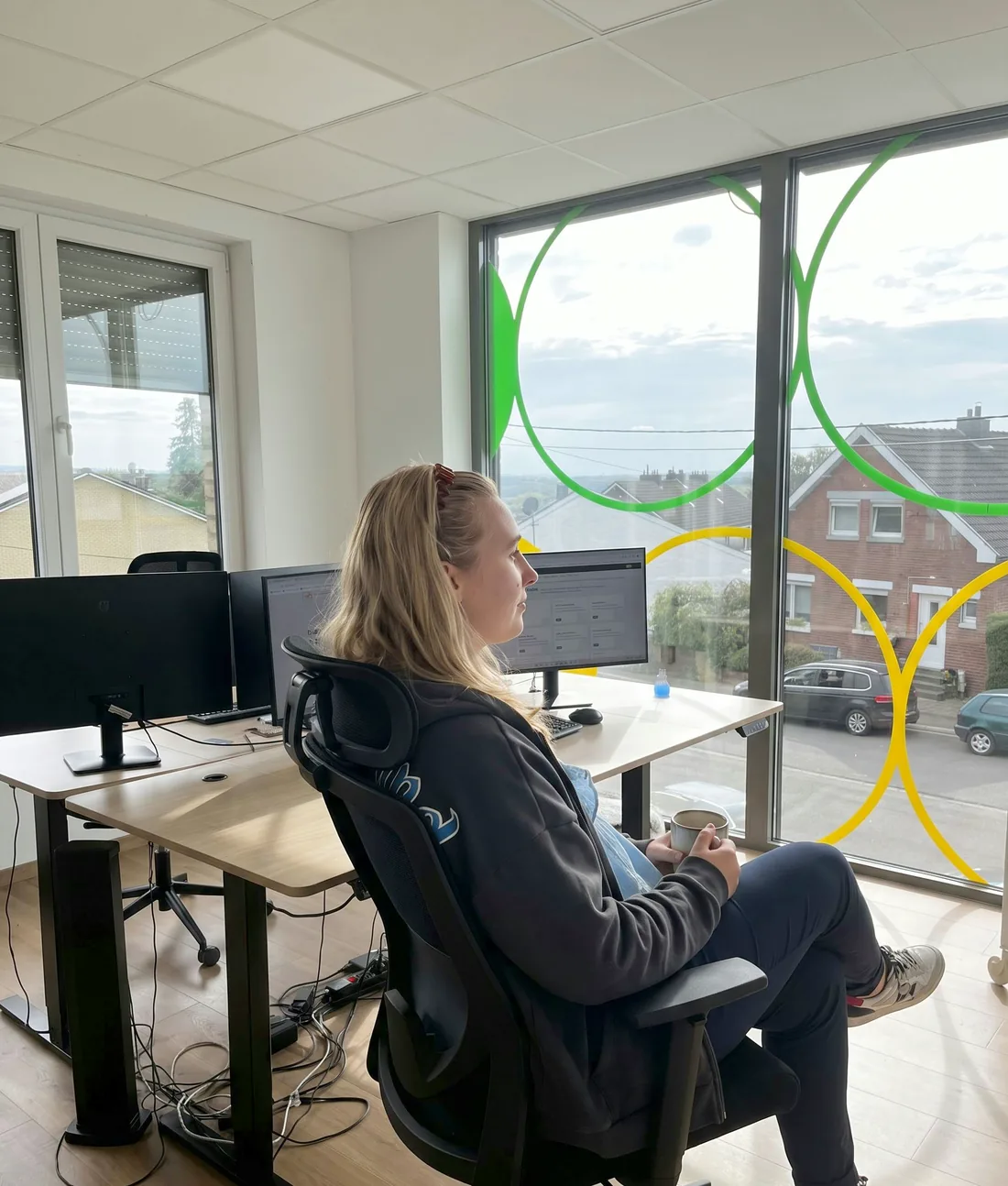 Eine Frau sitzt mit einer Tasse am Schreibtisch im Büro von Bits & Bots und schaut aus dem Fenster